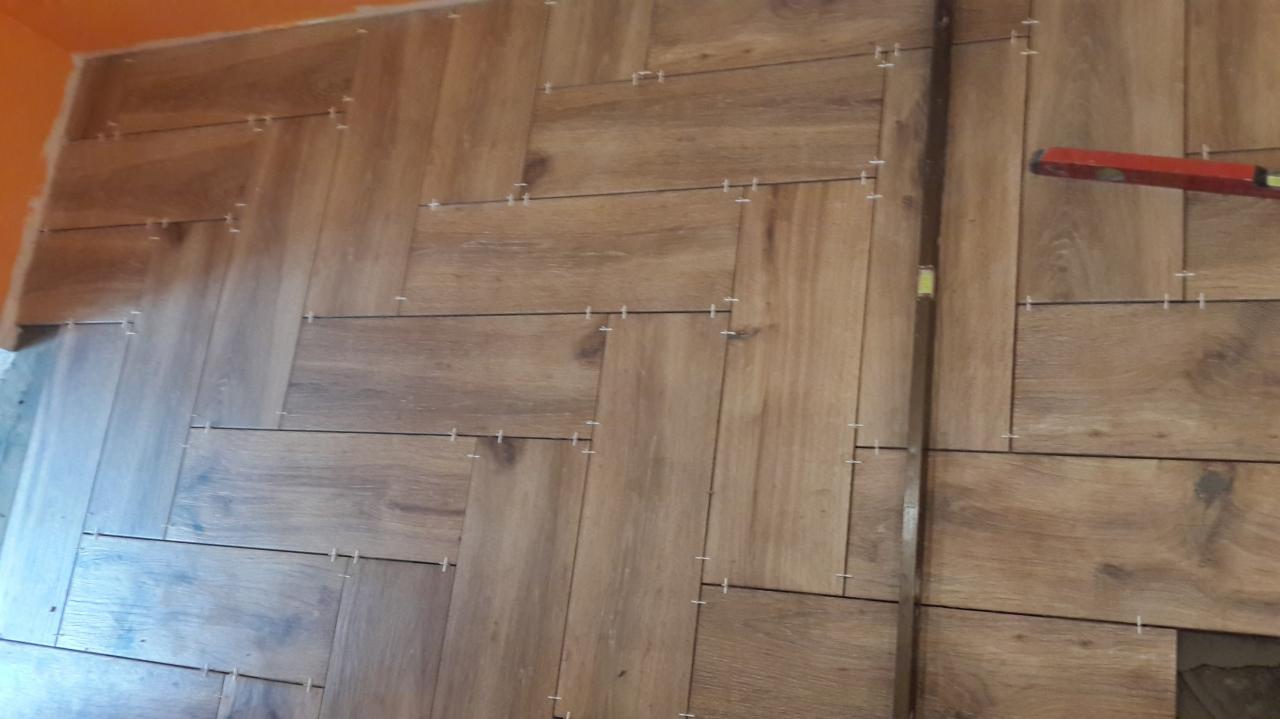 Układanie brązowych płytek imitujących drewno na ścianie w jodełkę, z widocznymi krzyżykami dystansowymi i poziomicą przyłożoną do pionowej listwy.