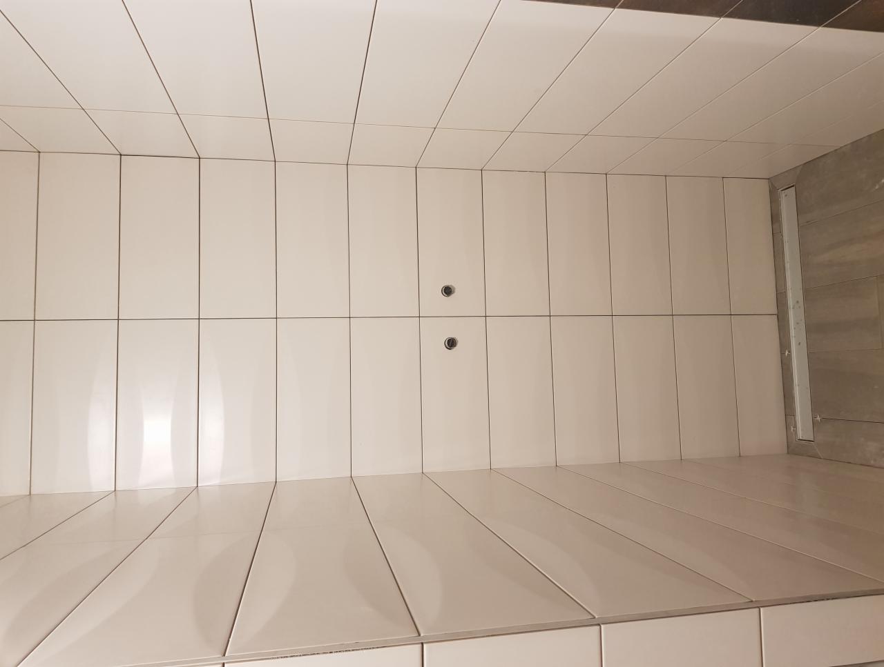 Wnętrze kabiny prysznicowej wyłożonej białymi, prostokątnymi płytkami ceramicznymi, z widocznymi dwoma otworami na baterię oraz odpływem liniowym przy ścianie z szarymi płytkami.