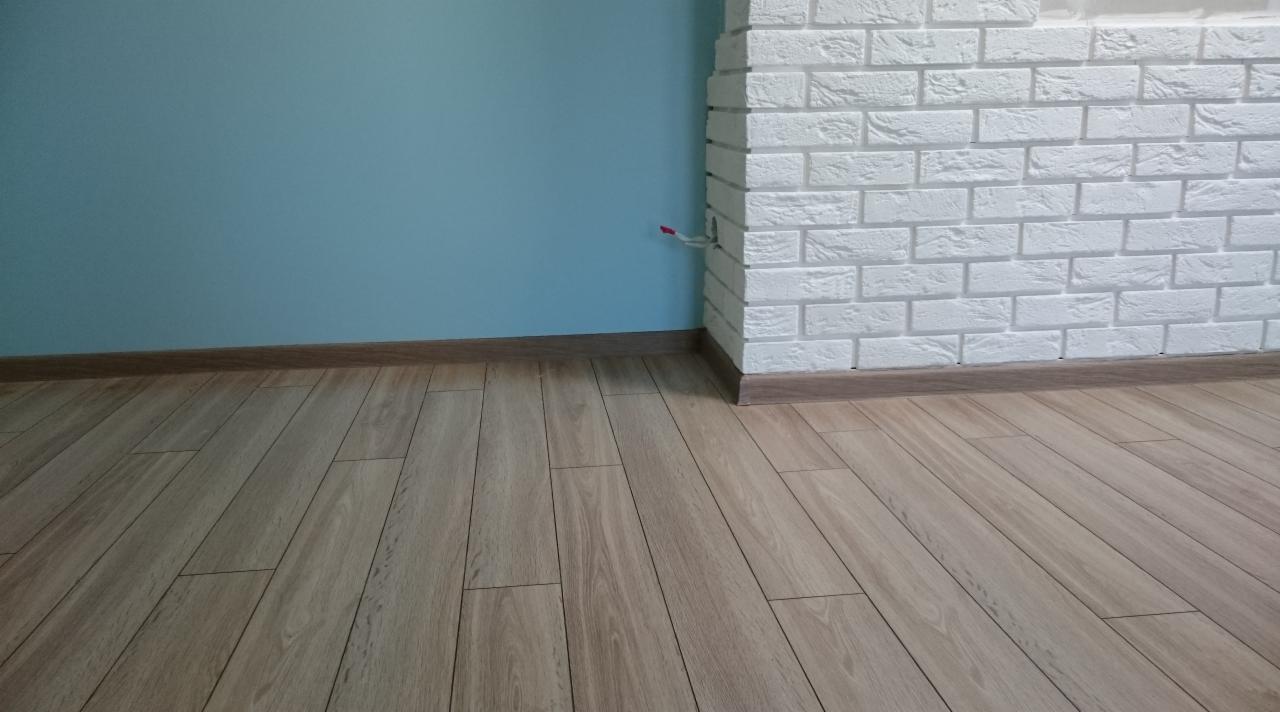 Fragment wnętrza z panelami podłogowymi o wzorze drewna, jasnoniebieską ścianą i fragmentem ściany wyłożonej białymi cegłami, wykończonej brązową listwą przypodłogową. Widoczny wystający przewód.