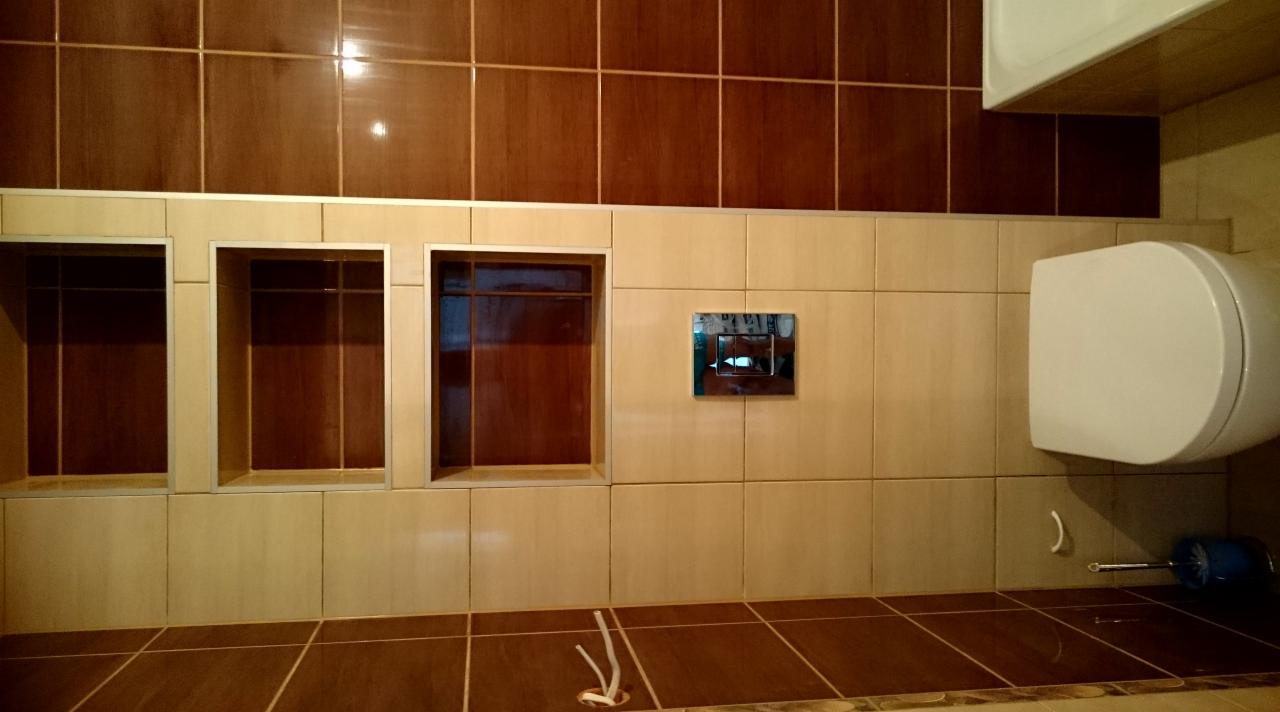 Wykończona łazienka z beżowo-brązowymi płytkami, trzema wnękami w ścianie, wiszącą toaletą i chromowanym przyciskiem spłuczki. Na podłodze leżą białe przewody.