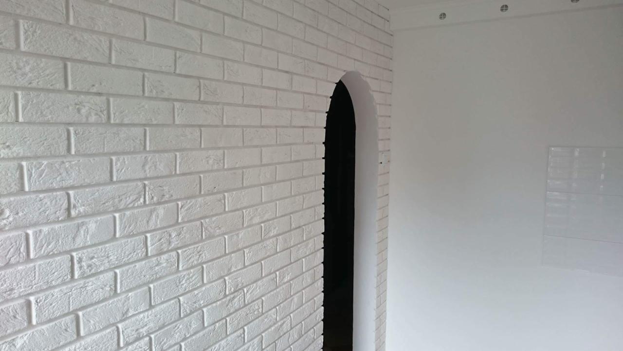 Ściana wykończona białymi płytkami imitującymi cegłę, z łukowatym przejściem do sąsiedniego pomieszczenia, w tle fragment białej ściany z widocznymi elementami wentylacji sufitowej.