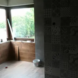 Wnętrze w trakcie remontu: ułożona podłoga z paneli imitujących drewno, ściana wyłożona dekoracyjnymi płytkami, widoczne materiały budowlane, surowa żarówka zwisająca z sufitu.