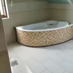 Wanna narożna obudowana mozaiką w łazience na poddaszu, białe płytki podłogowe, sufit z płyt karton gips.