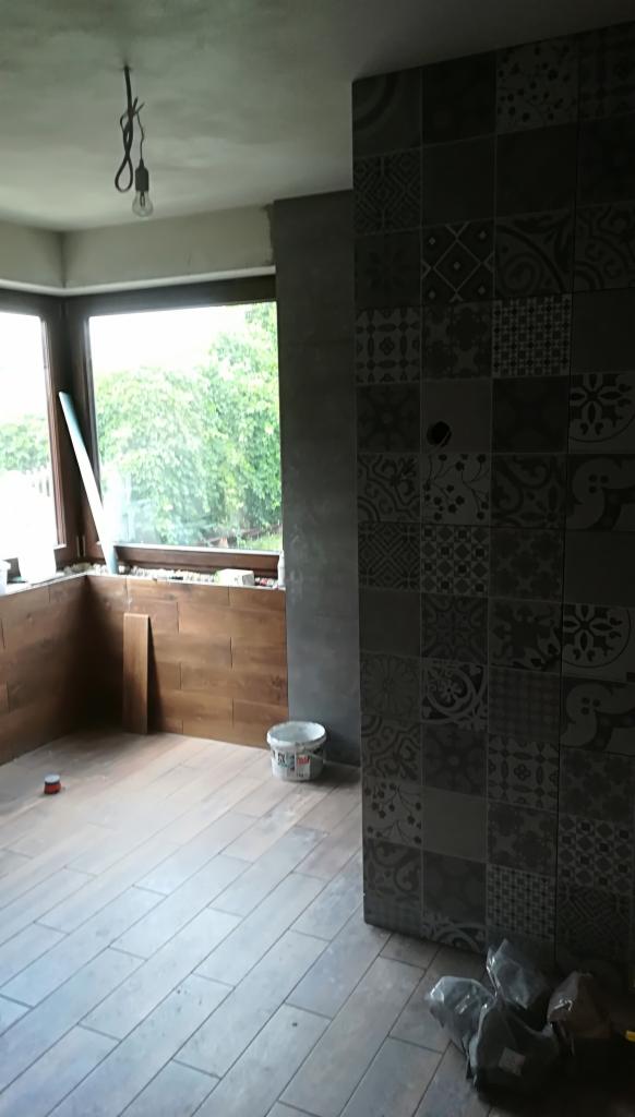 Wnętrze w trakcie remontu: ułożona podłoga z paneli imitujących drewno, ściana wyłożona dekoracyjnymi płytkami, widoczne materiały budowlane, surowa żarówka zwisająca z sufitu.