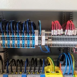 Panel elektryczny z licznymi kablami w różnych kolorach (niebieski, czarny, czerwony, żółto-zielony) podłączonymi do listew zaciskowych z numeracją i oznaczeniami.