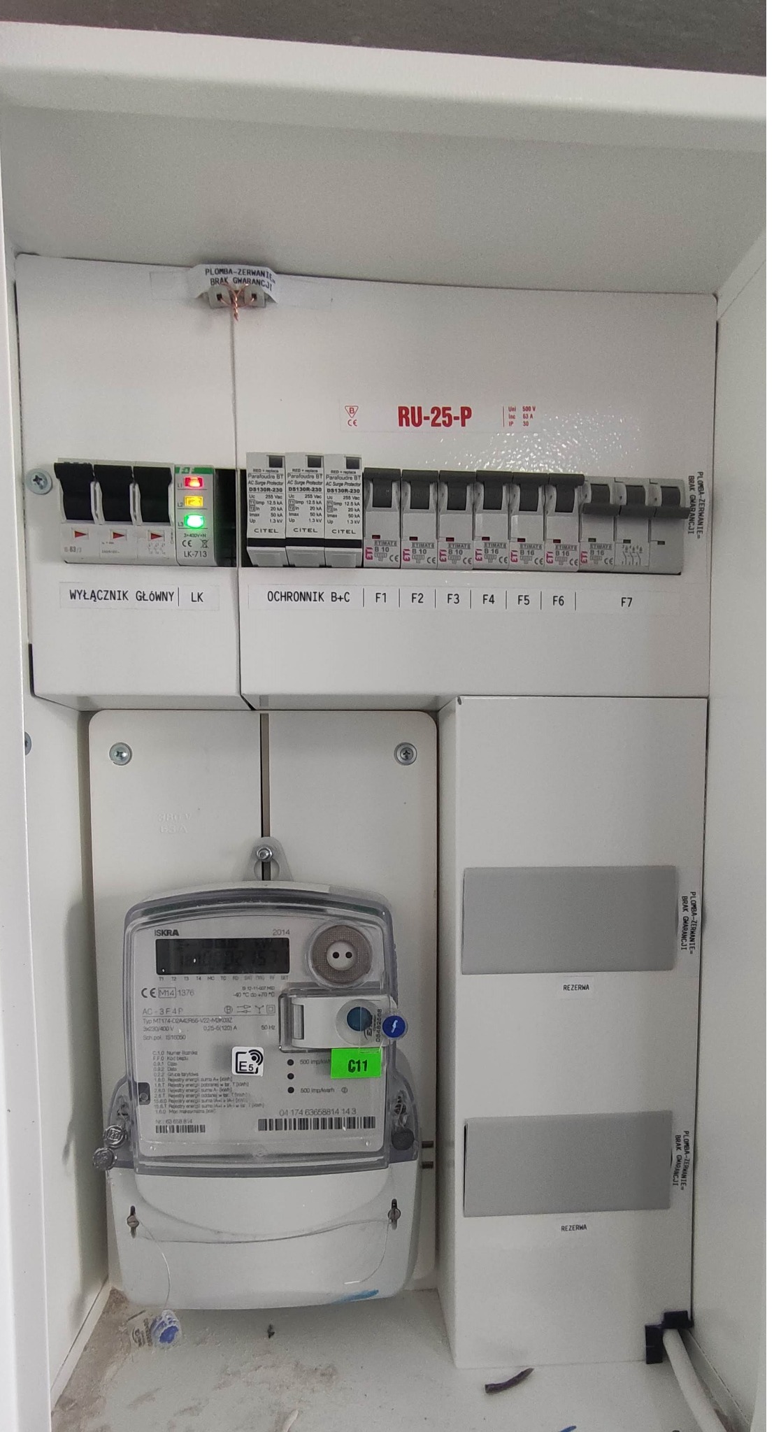 Skrzynka elektryczna z licznikiem energii, wyłącznikiem głównym, bezpiecznikami i ogranicznikiem przepięć, widoczne oznaczenia i plomby.