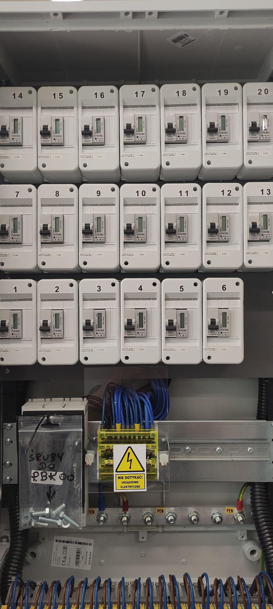 Rozdzielnica elektryczna z zamontowanymi bezpiecznikami, widoczne okablowanie, listwa zaciskowa z ostrzeżeniem o napięciu i woreczek ze śrubami do PBK00