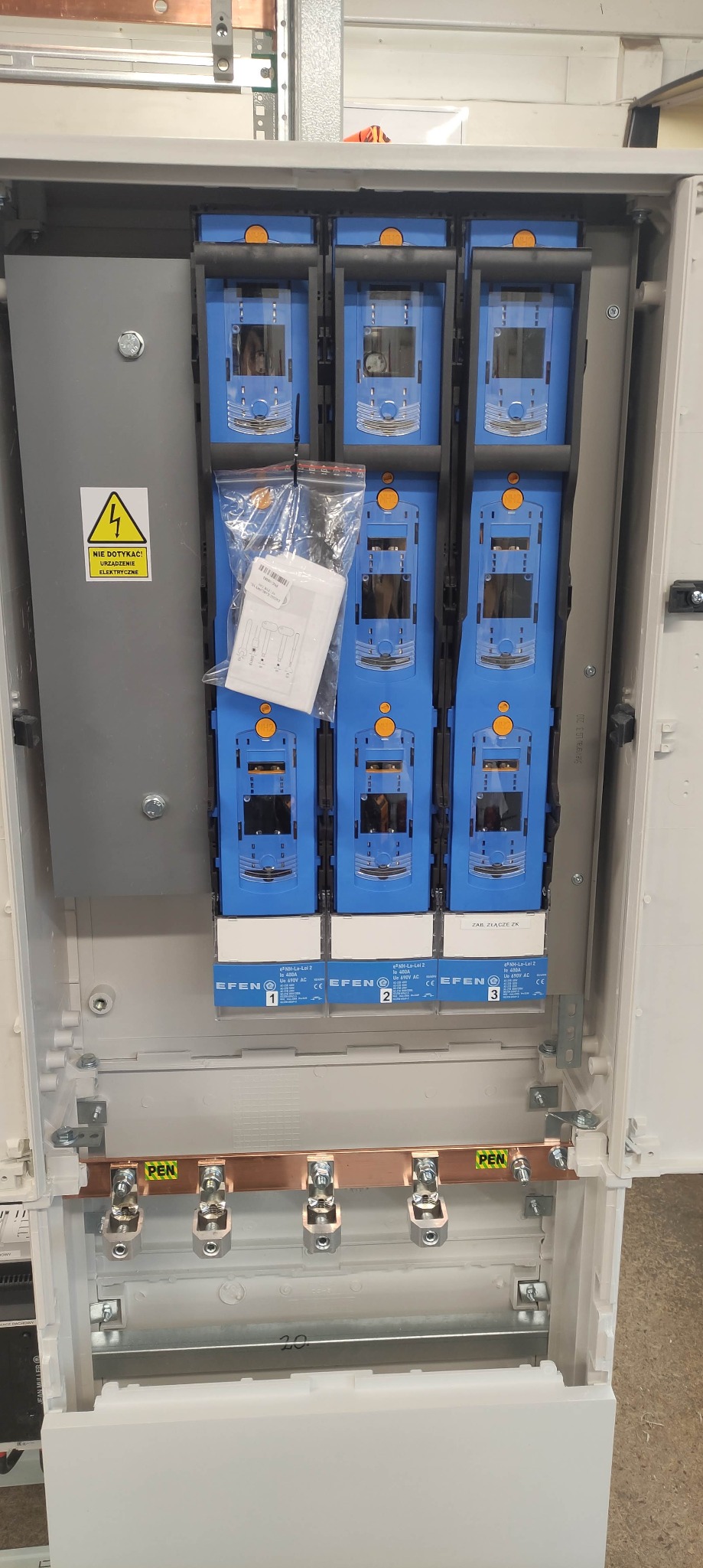 Rozdzielnia elektryczna z zamontowanymi bezpiecznikami EFEN, widoczna miedziana szyna PEN, naklejka ostrzegawcza 'Nie dotykać! Urządzenie elektryczne' oraz instrukcja w folii.
