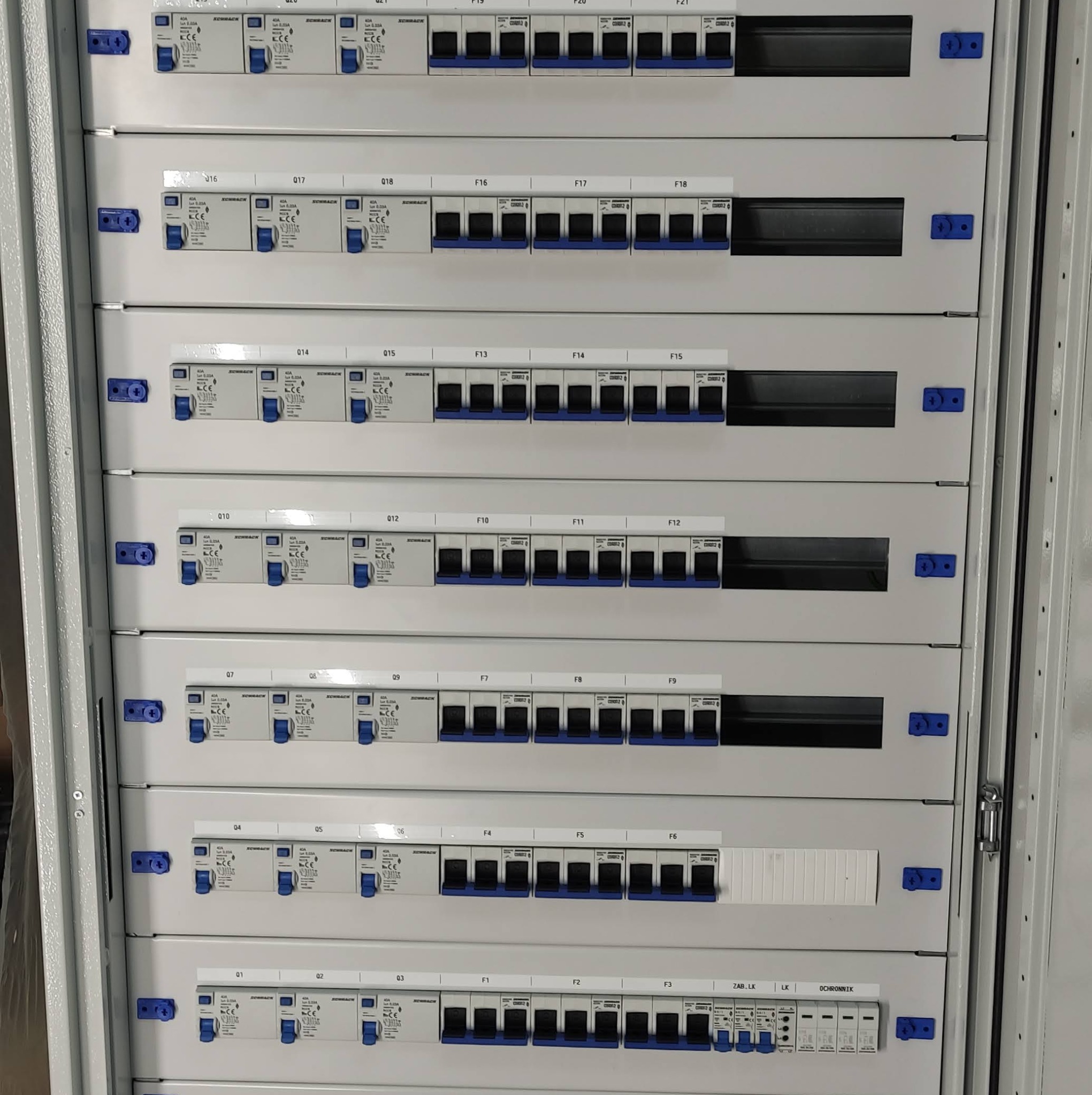 Szczegółowy widok rozdzielnicy elektrycznej z wieloma zabezpieczeniami nadprądowymi i różnicowoprądowymi, zamontowanymi w szarym metalowym panelu z niebieskimi elementami mocującymi.