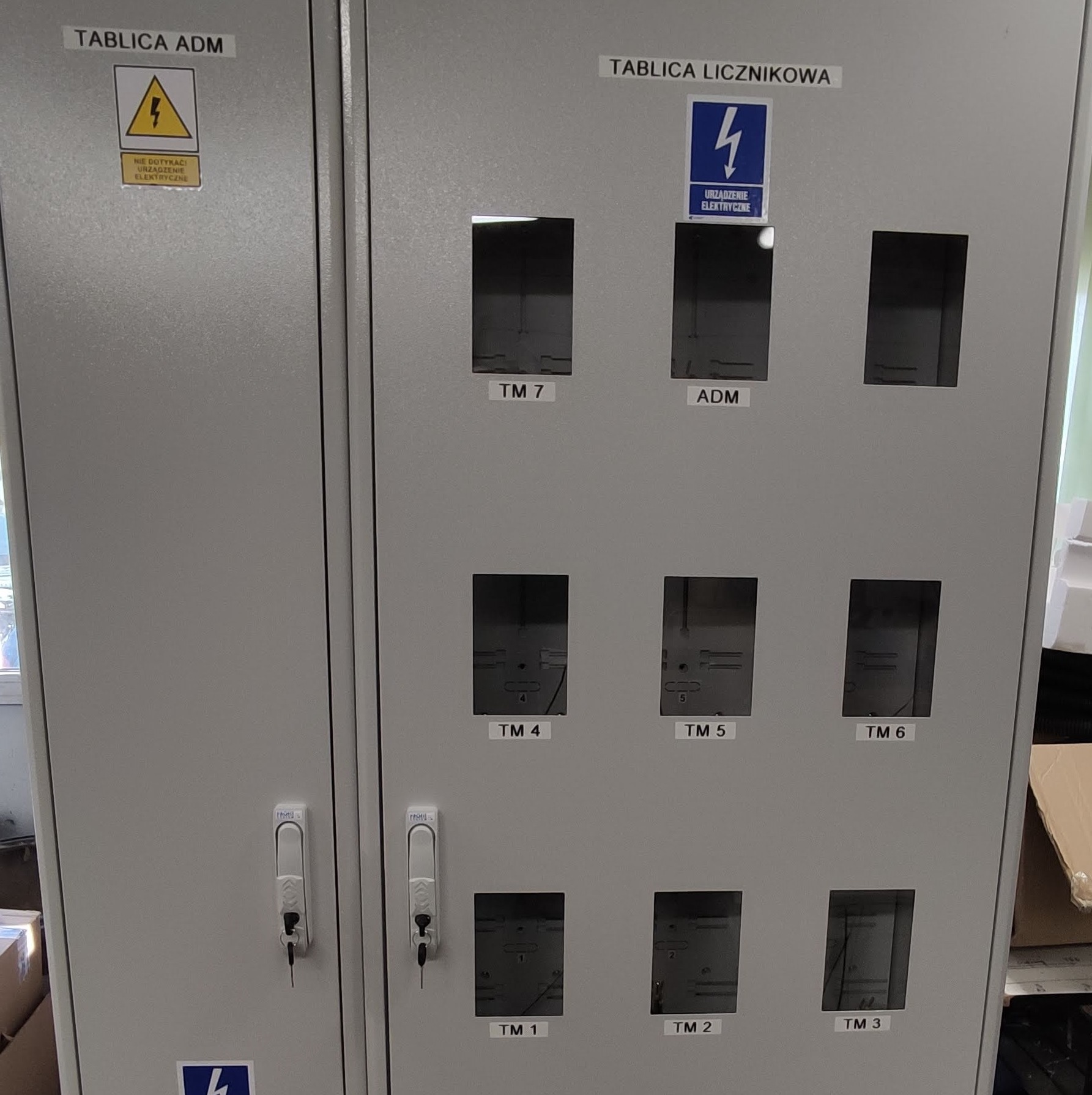 Szara tablica licznikowa z otwieranymi drzwiczkami, widoczne oznaczenia TM 1-7 oraz ADM, naklejka ostrzegawcza o urządzeniu elektrycznym.
