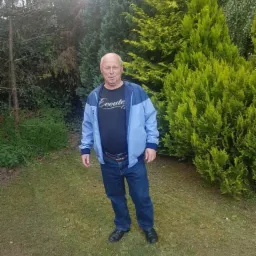 Mężczyzna w niebieskiej kurtce stoi na trawniku przed zielonymi drzewami iglastymi w słoneczny dzień. Widoczne jeansy i czarne buty.