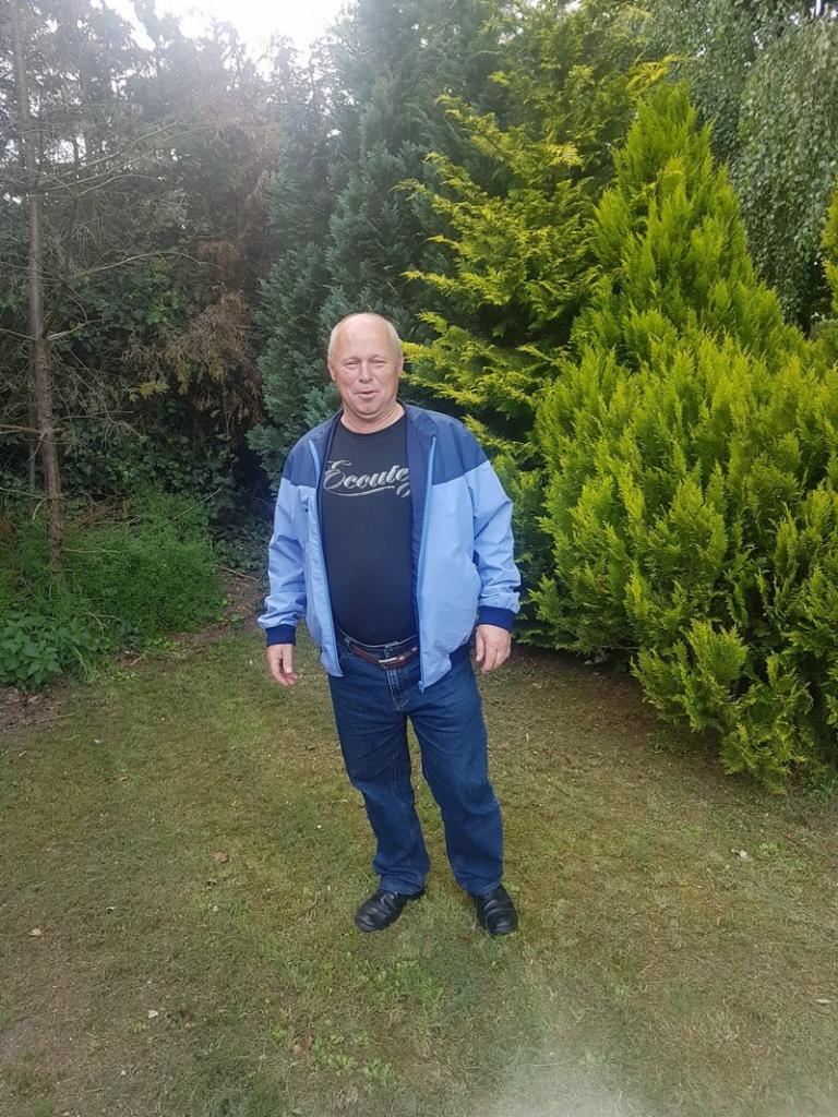 Mężczyzna w niebieskiej kurtce stoi na trawniku przed zielonymi drzewami iglastymi w słoneczny dzień. Widoczne jeansy i czarne buty.