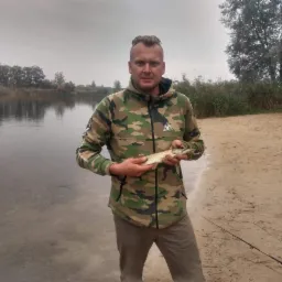 Mężczyzna w bluzie moro trzyma w rękach złowioną rybę (szczupak) na tle jeziora i piaszczystej plaży. W pobliżu wędka.