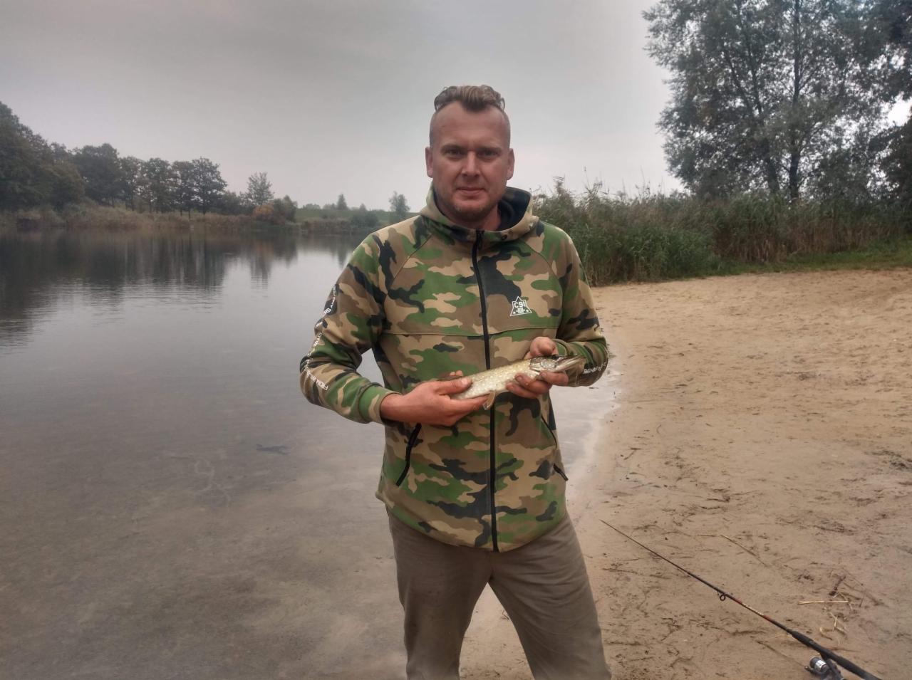 Mężczyzna w bluzie moro trzyma w rękach złowioną rybę (szczupak) na tle jeziora i piaszczystej plaży. W pobliżu wędka.
