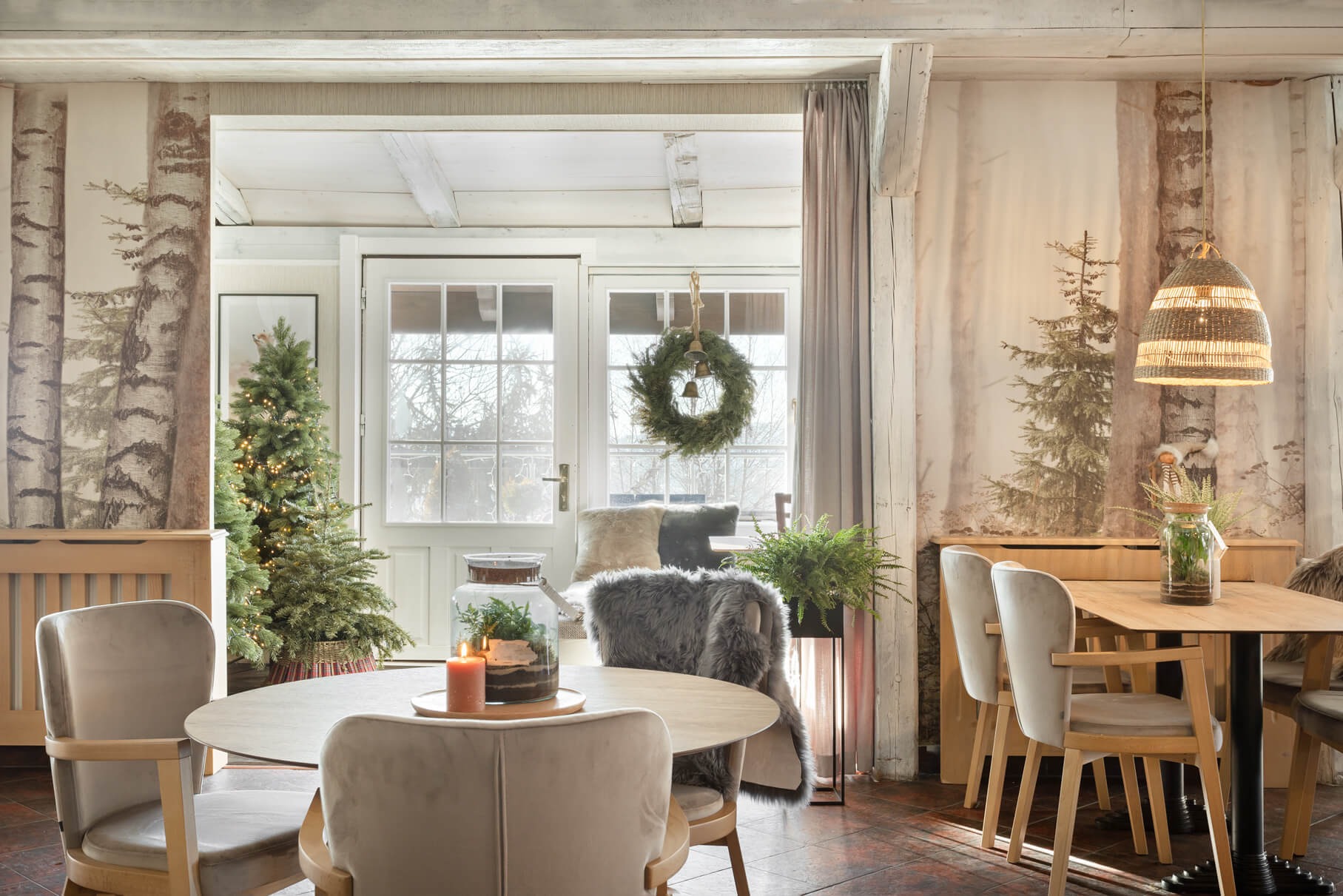 Jadalnia w stylu rustykalnym z motywem leśnym: tapeta z brzozami, choinka, wieniec świąteczny na drzwiach, drewniane meble, beżowe krzesła z weluru, świeca na stole i lampion wiszący nad nim.