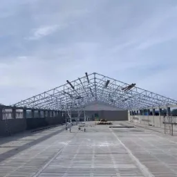 Konstrukcja stalowa hali w trakcie budowy, widoczne rusztowania, betonowa podstawa i elementy dachu, jasne niebo w tle.
