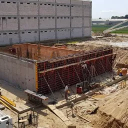 Wykop pod budynek przemysłowy z częściowo wylanymi betonowymi ścianami, szalunkami i zbrojeniami, widoczne dwie betoniarki z pompami, w tle budynek w trakcie budowy.