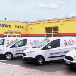 Flota białych samochodów dostawczych z logo Centrum Hurtowego Pasz zaparkowana przed żółtym budynkiem hurtowni.