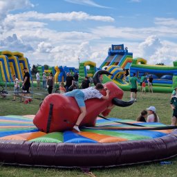 Agencja DK - Chłopiec próbuje utrzymać się na dmuchanym byku rodeo na tle dmuchanych zamków i zjeżdżalni na imprezie plenerowej.