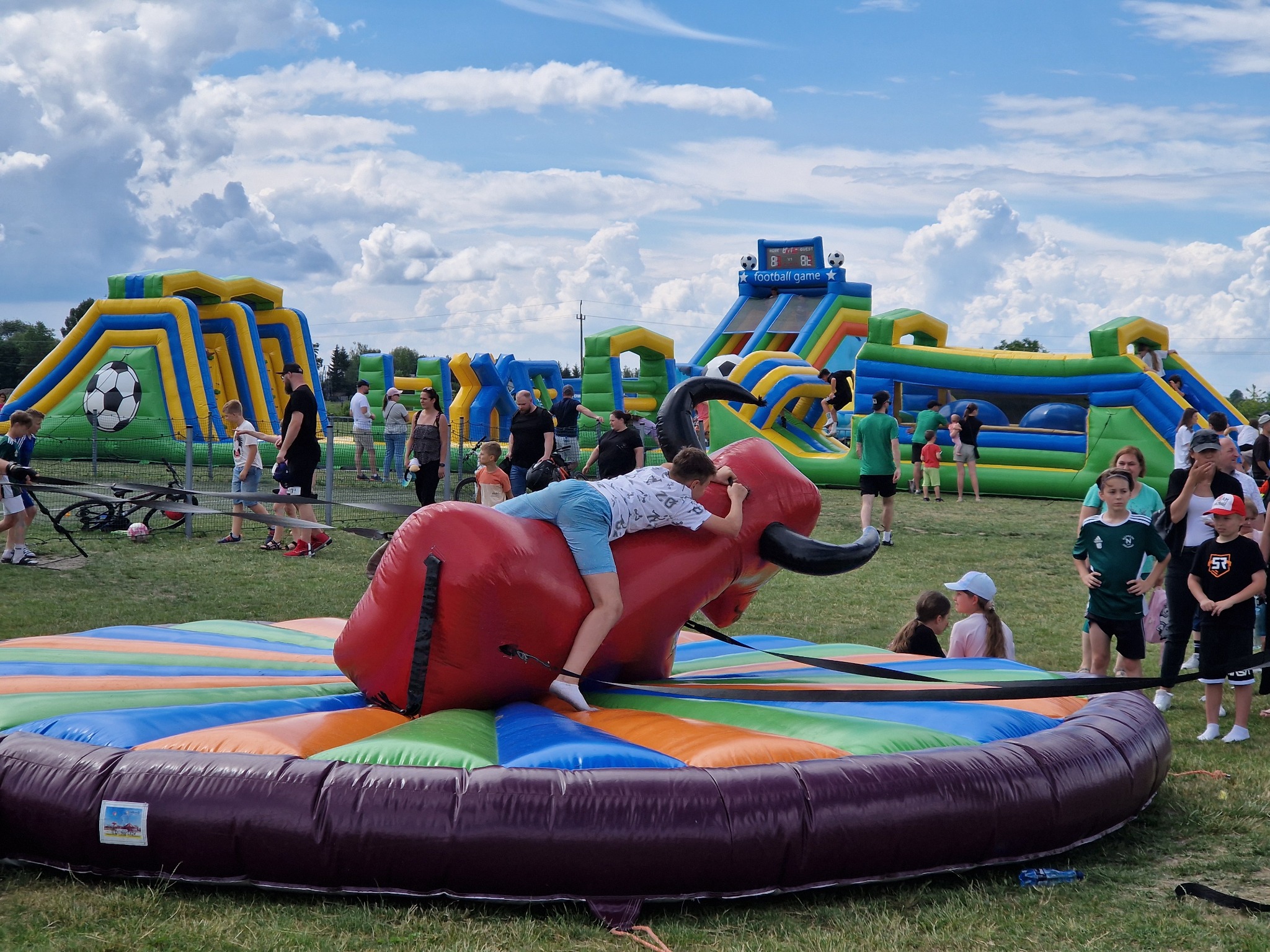 Chłopiec próbuje utrzymać się na dmuchanym byku rodeo na tle dmuchanych zamków i zjeżdżalni na imprezie plenerowej.