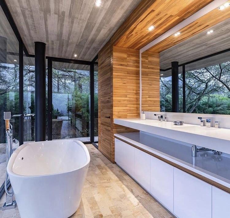 Nowoczesna łazienka z dużą wanną wolnostojącą, przeszkloną ścianą z widokiem na zieleń, drewnianą boazerią na ścianie i podwójną umywalką z białym blatem.