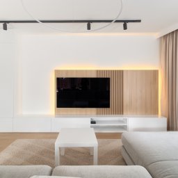 JLT Wykończenia wnętrz Dariusz Kóska - Nowoczesny salon z zabudową TV z podświetleniem LED, minimalistyczna biała szafka RTV i regał z drewnianymi wnękami. Beżowe zasłony i dywan.