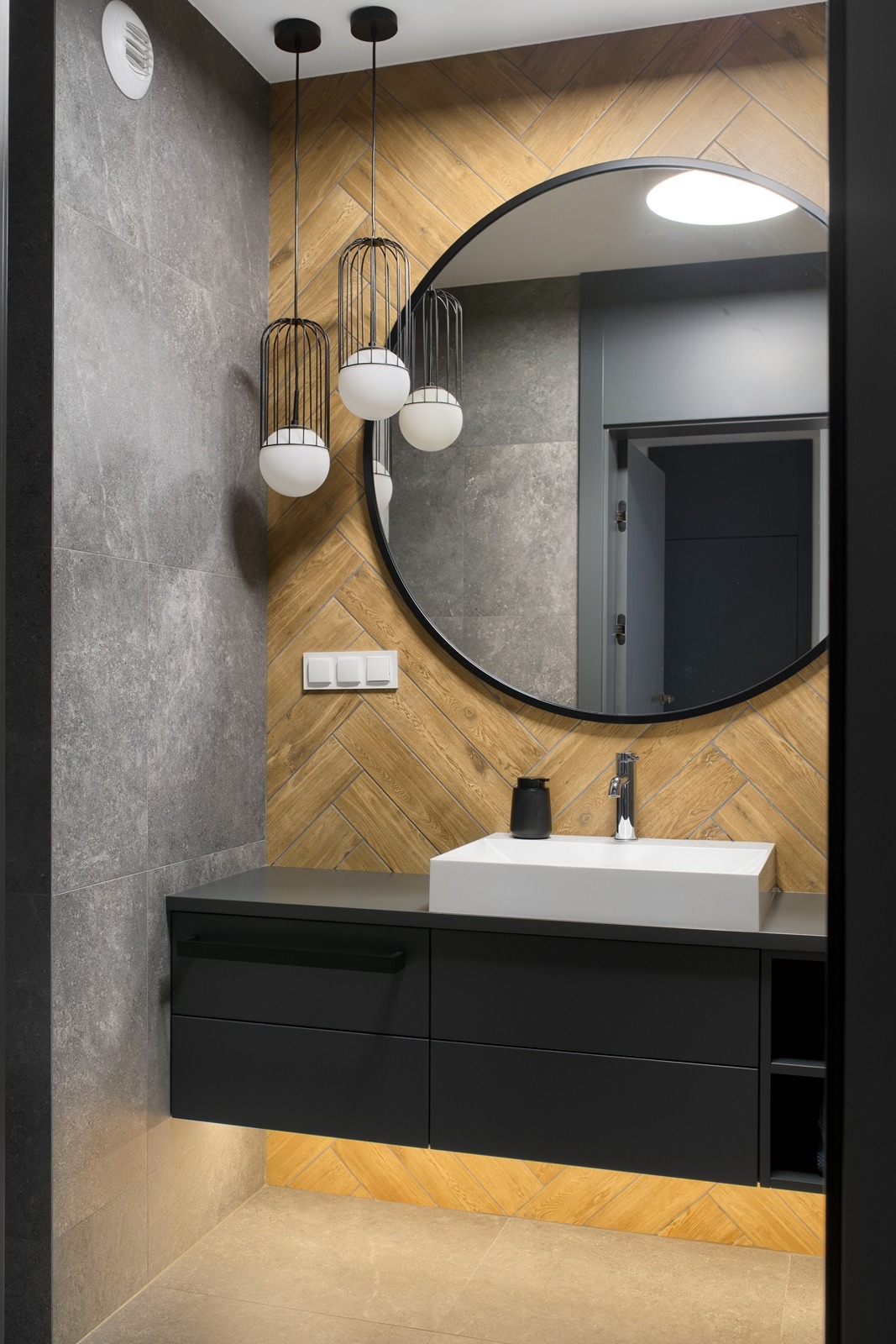 Nowoczesna łazienka w Dopiewie z szarą płytką, drewnianą mozaiką i okrągłym lustrem. Umywalka nablatowa i czarna szafka z podświetleniem LED.