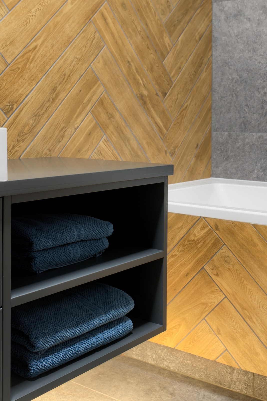 Wykończona łazienka z drewnopodobną glazurą ułożoną w jodełkę, szarą szafką z ręcznikami i fragmentem wanny. Nowoczesne wzornictwo i dbałość o detale.