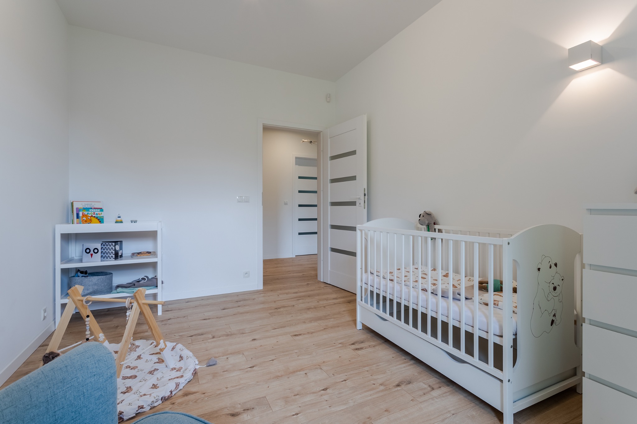 Jasny pokój dziecięcy z białym łóżeczkiem, regałem i drewnianą podłogą. W tle otwarte drzwi z poziomymi przeszkleniami i minimalistyczna lampa ścienna.