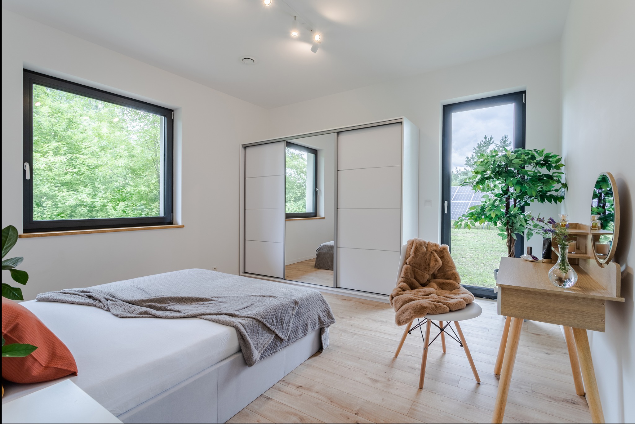Jasna sypialnia z drewnianą podłogą, białymi ścianami i szafą przesuwną z lustrem. Widok na zieleń za oknem. Minimalistyczny wystrój z łóżkiem i toaletką.