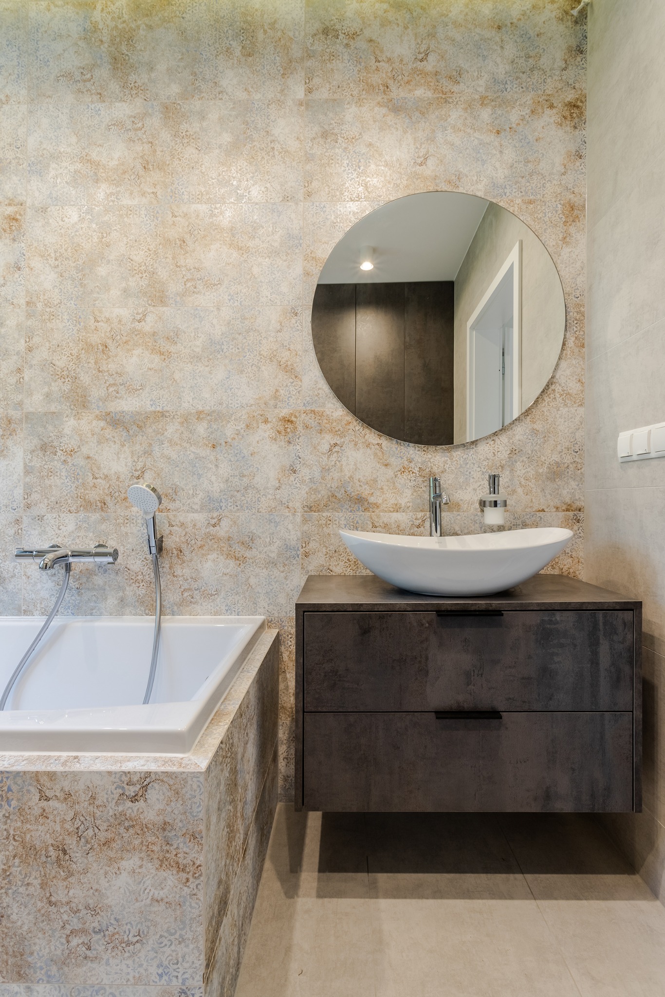 Wykończona łazienka z wanną obudowaną płytkami wzorzystymi, umywalką nablatową na szafce z szufladami i okrągłym lustrem na ścianie.