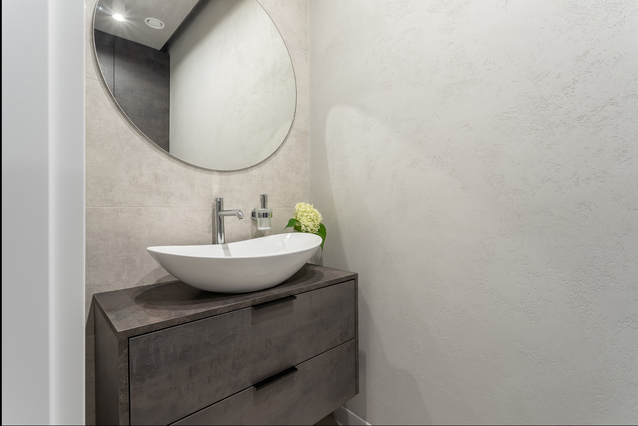Nowoczesna łazienka z owalną umywalką na szafce z szufladami, okrągłe lustro na tle ściany z dekoracyjną fakturą. Minimalistyczny design, stonowana kolorystyka.