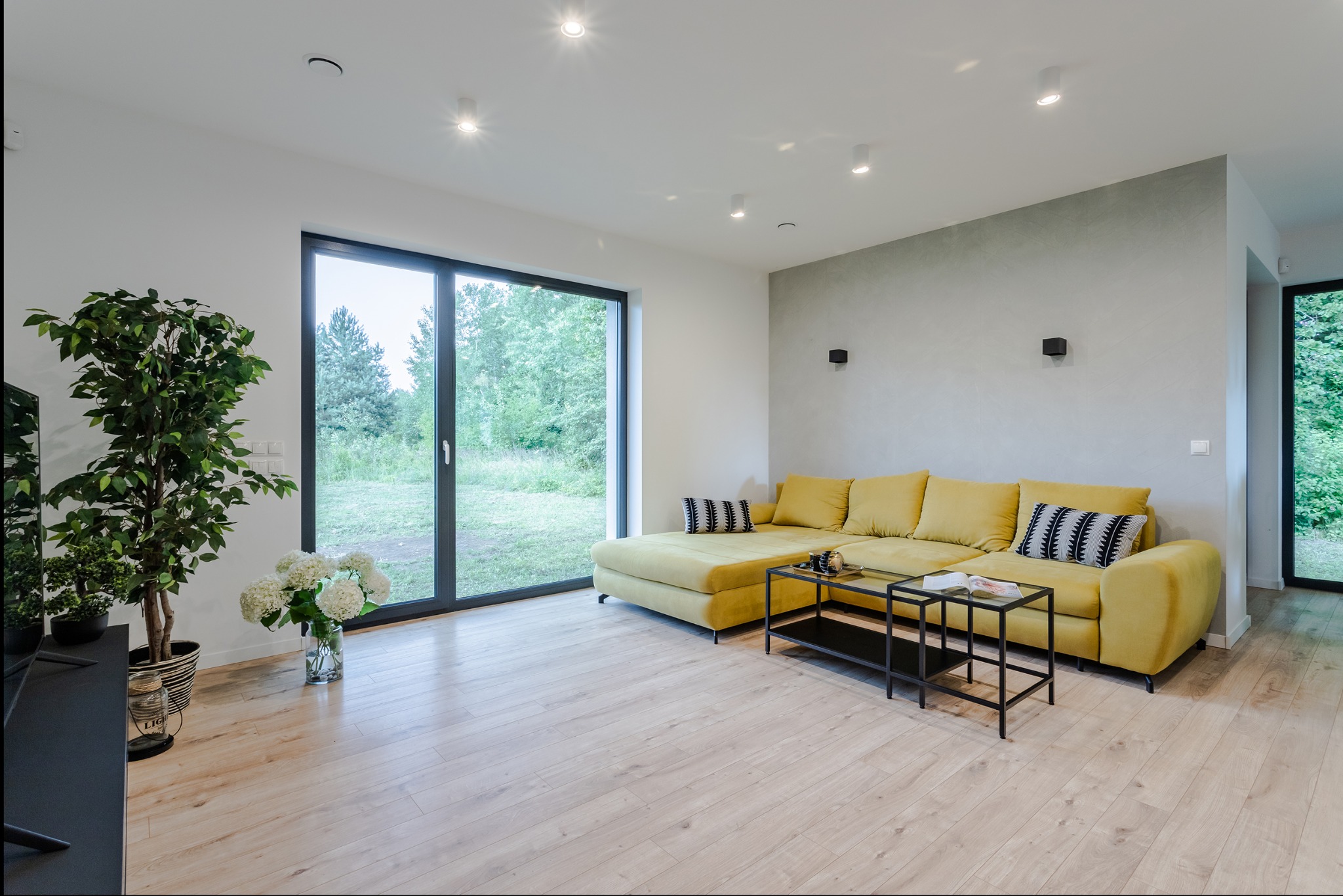 Jasny salon z żółtą kanapą narożną, drewnianą podłogą i dużym oknem z widokiem na zieleń. Ściana za kanapą wykończona w odcieniach szarości.