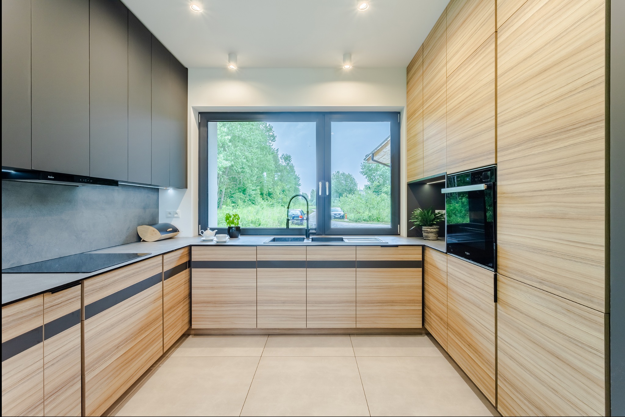 Nowoczesna kuchnia w odcieniach szarości i drewna z dużym oknem. Widok na zieleń za oknem. Minimalistyczny design, oświetlenie punktowe.