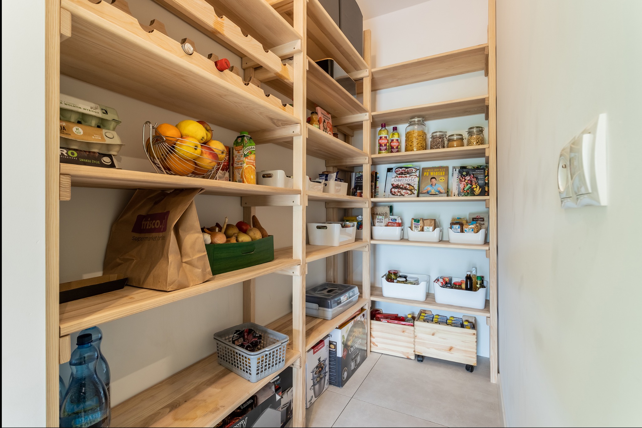 Jasna, drewniana spiżarnia z półkami pełnymi produktów spożywczych, od jaj i owoców po słoiki z przyprawami i książki kucharskie. Widoczna instalacja elektryczna.