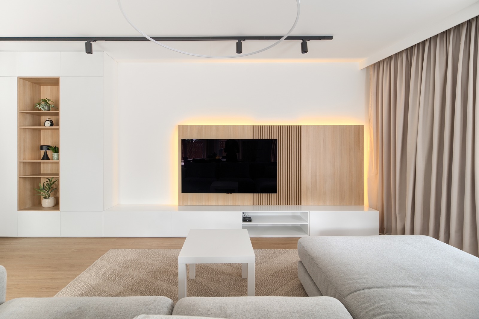 Nowoczesny salon z zabudową TV z podświetleniem LED, minimalistyczna biała szafka RTV i regał z drewnianymi wnękami. Beżowe zasłony i dywan.