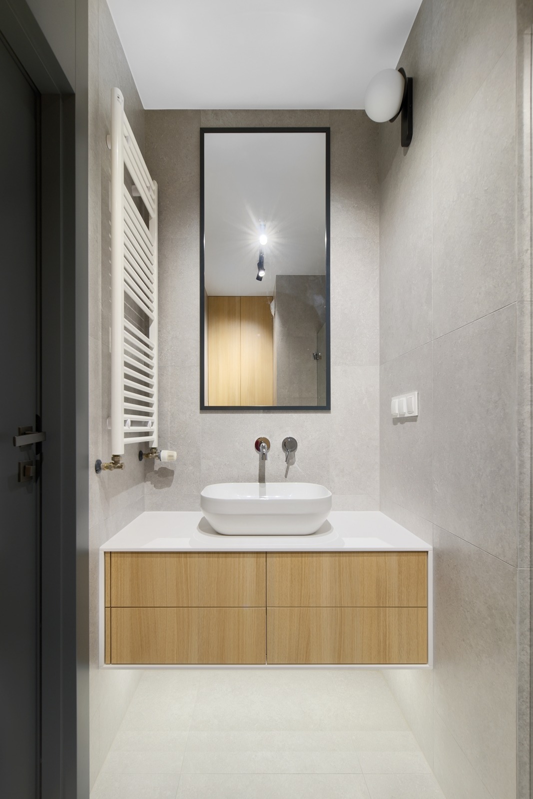 Nowoczesna łazienka z umywalką nablatową, drewnianą szafką pod umywalką, szarymi płytkami i dużym lustrem w czarnej ramie. Minimalistyczny design.