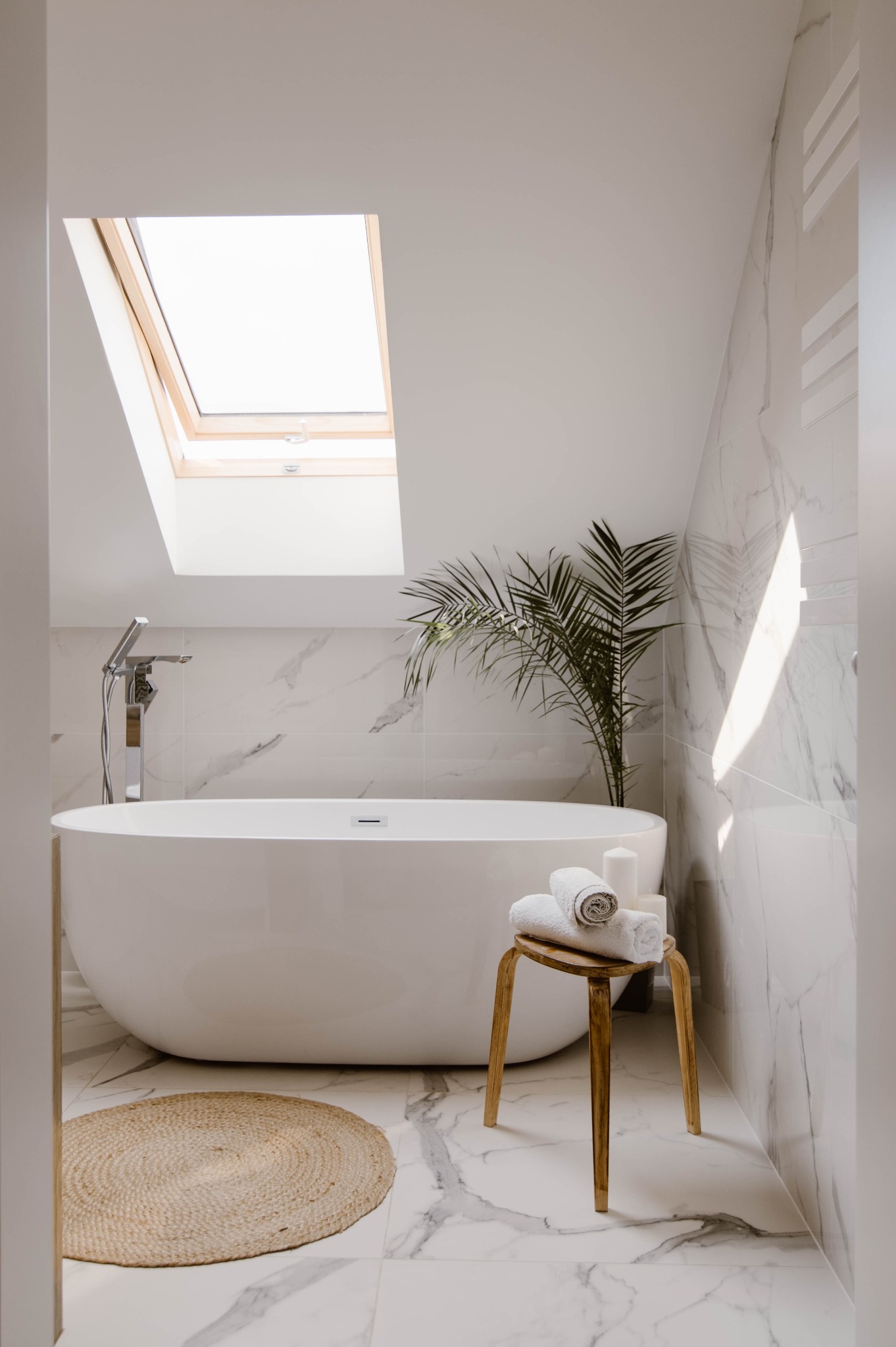 Nowoczesna łazienka na poddaszu z marmurowymi płytkami, wolnostojącą wanną, oknem dachowym i elementami dekoracyjnymi w stylu skandynawskim.