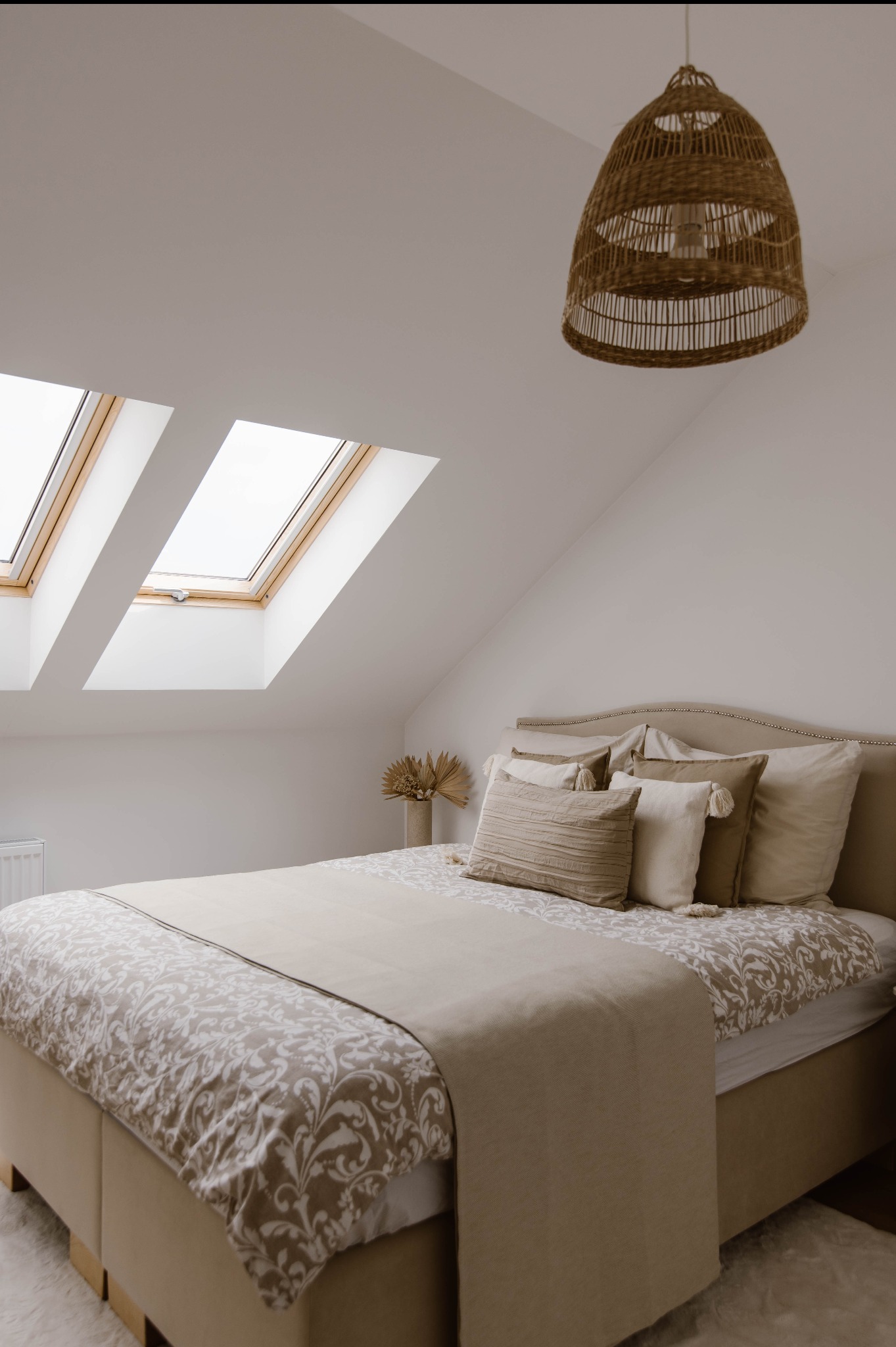 Beżowa sypialnia na poddaszu z dwoma oknami dachowymi. Wnętrze doświetlone przez wiklinową lampę. Na łóżku dekoracyjne poduszki i narzuta z motywem roślinnym.
