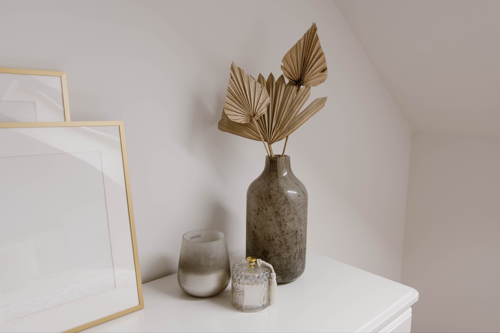 Dekoracyjny kącik: złote ramy, szklany świecznik, marmurkowy wazon z suszem i kryształowy dyfuzor zapachowy na białej komodzie w jasnym wnętrzu.