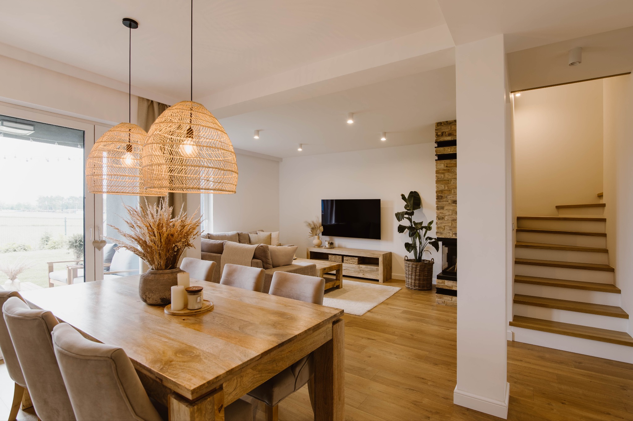 Jasne wnętrze domu z drewnianym stołem, krzesłami i ozdobnymi lampami wiszącymi. W tle salon z telewizorem i schody na piętro. Subtelne oświetlenie.