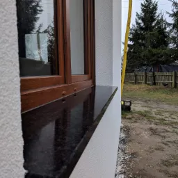 Czarny, błyszczący parapet zewnętrzny z konglomeratu, zamontowany pod oknem z drewnianą ramą; widoczne drzewa w tle.