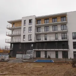 Nowoczesny budynek apartamentowy z balkonami w trakcie budowy, widoczny teren budowy z błotem i materiałami budowlanymi, na dachu dwie osoby.