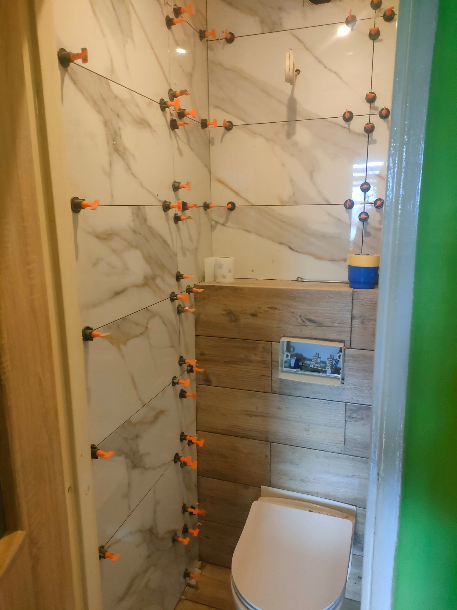 Łazienka w trakcie wykańczania, widoczne płytki imitujące marmur na ścianach z systemem poziomowania płytek, drewnopodobne płytki na zabudowie stelaża WC, przygotowane gniazdko elektryczne.