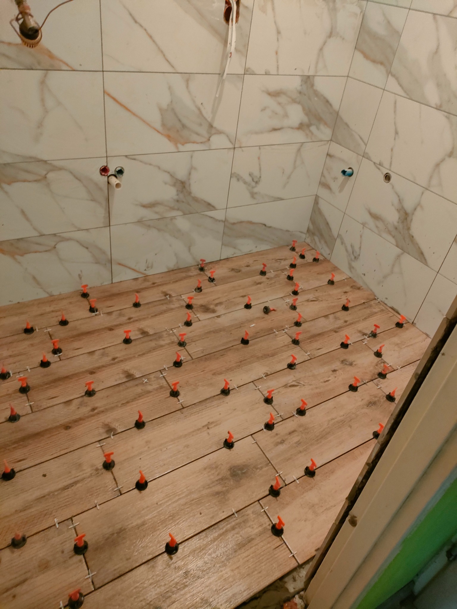 Podłoga w łazience w trakcie układania płytek imitujących drewno, z użyciem systemu poziomowania płytek z pomarańczowymi klinami, ściany wyłożone białymi płytkami z marmurkowym wzorem.