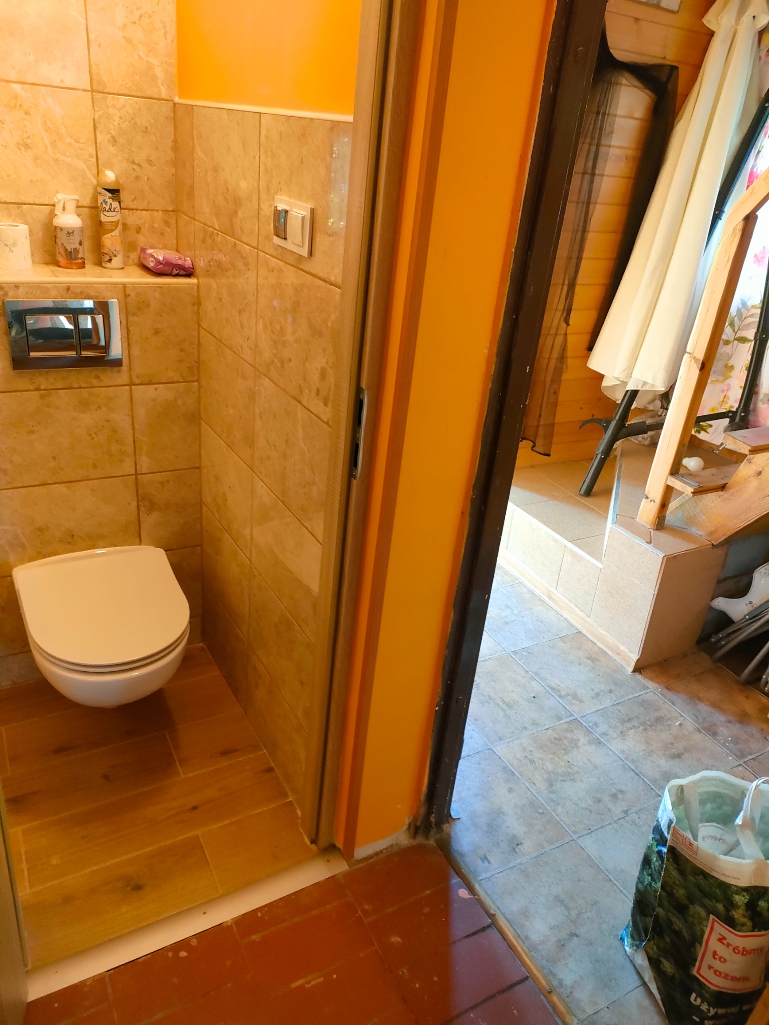 Ukończona łazienka z beżowymi płytkami imitującymi marmur, wisząca toaleta, drewnopodobna podłoga, widok zza otwartych drzwi na pomieszczenie z drewnianymi elementami i torbą z napisem 'Zróbmy to...