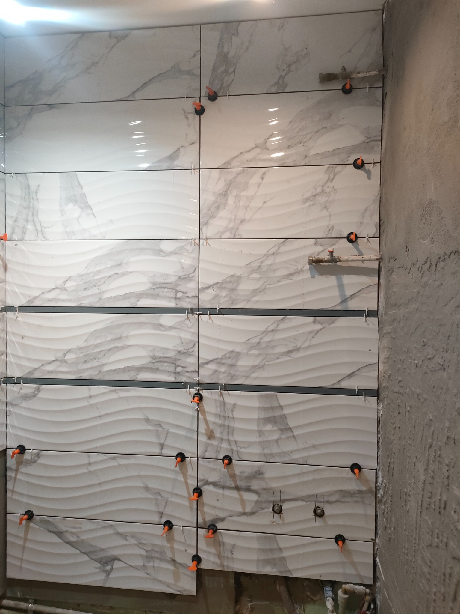 Montaż płytek imitujących marmur z falistym wzorem na ścianie w łazience, widoczne pomarańczowe klipsy poziomujące, surowy fragment ściany obok.