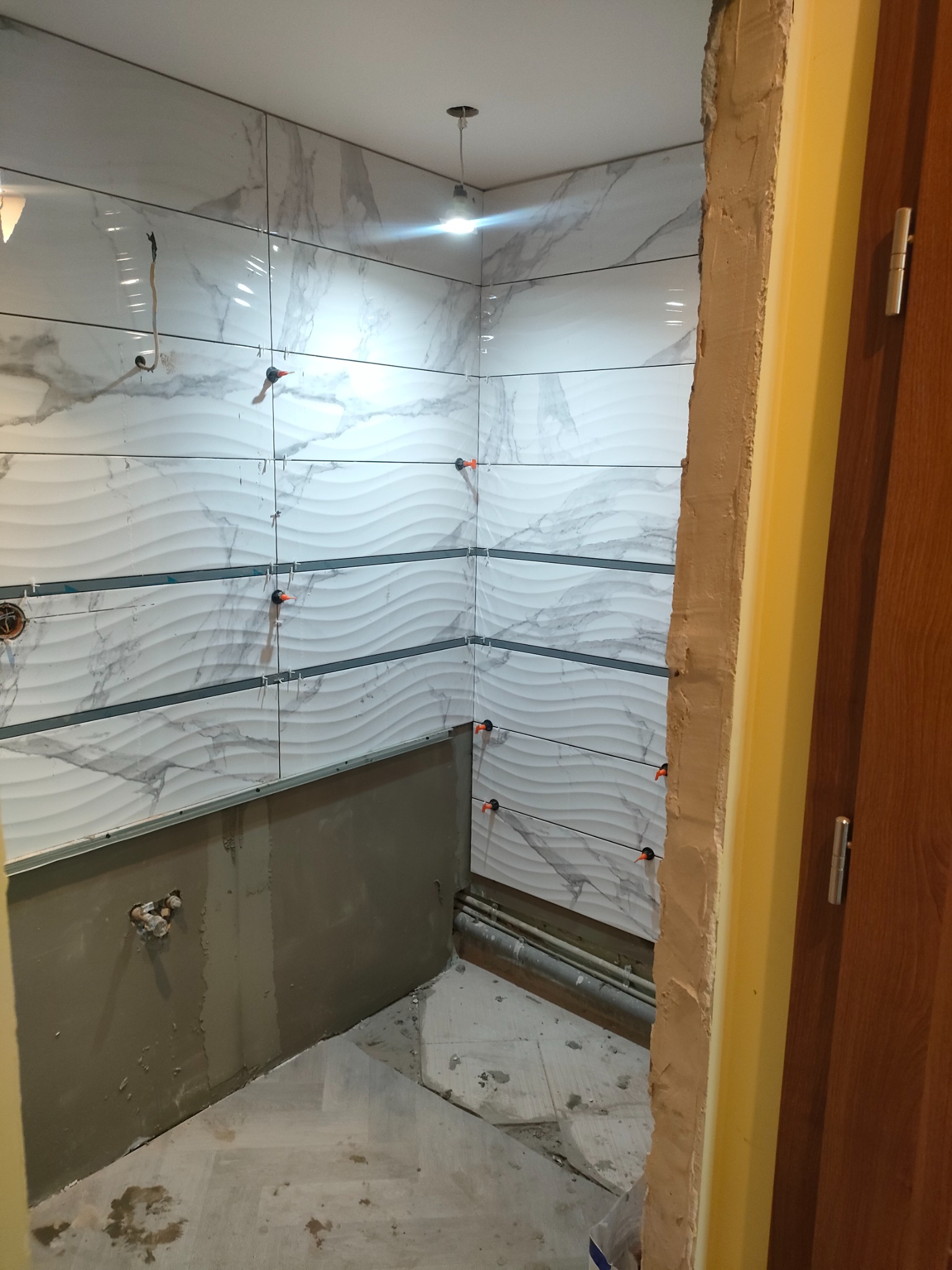 Łazienka w trakcie układania płytek z motywem fal, widoczne poziomice i krzyżyki dystansowe, częściowo wykończona ściana, surowy stan dolnej części ściany i podłogi.