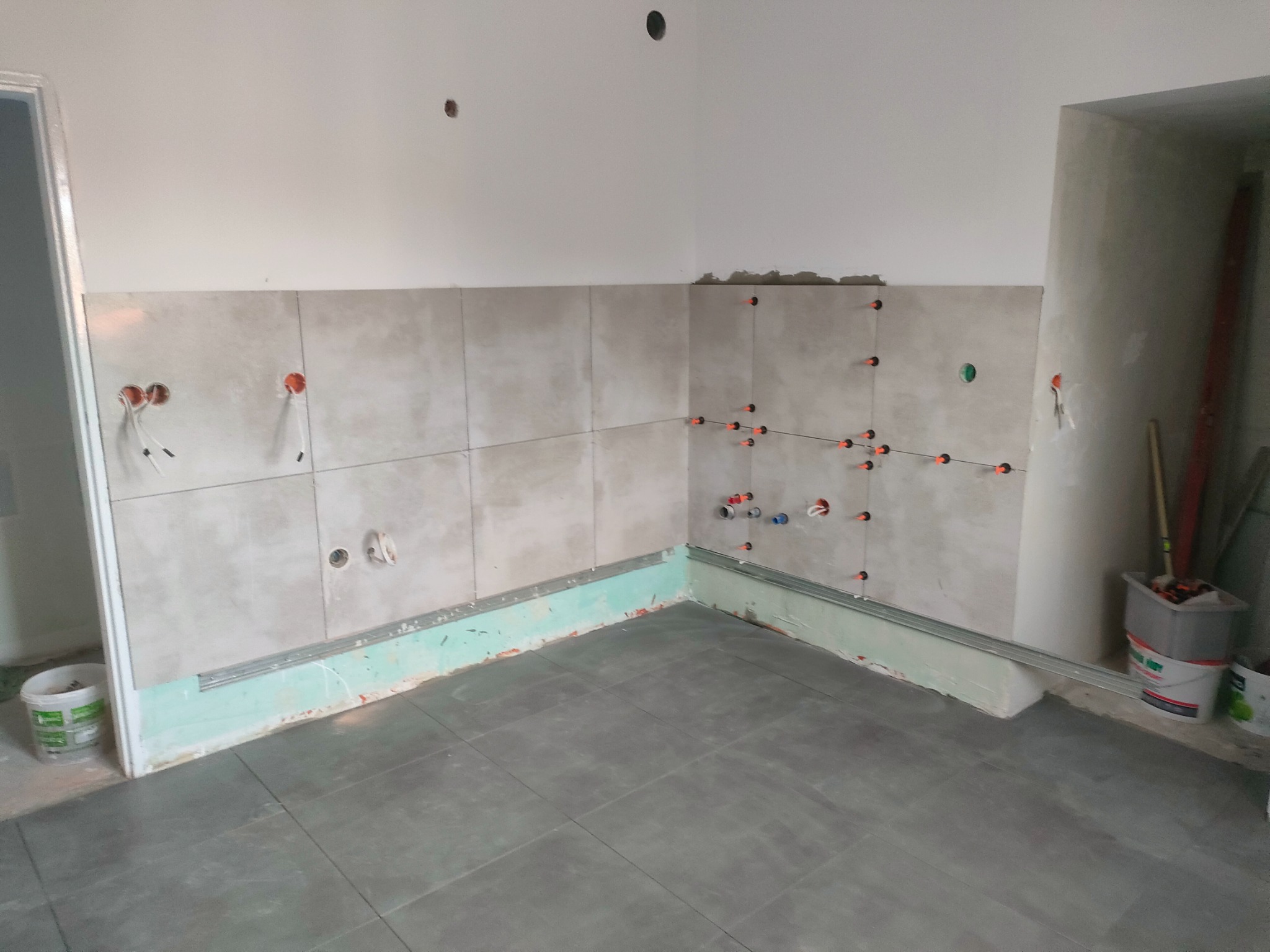 Wnętrze łazienki w trakcie remontu: ułożone szare płytki podłogowe, częściowo wyłożona ściana płytkami z krzyżykami dystansowymi, widoczne instalacje elektryczne i hydrauliczne, oraz gruntowanie...