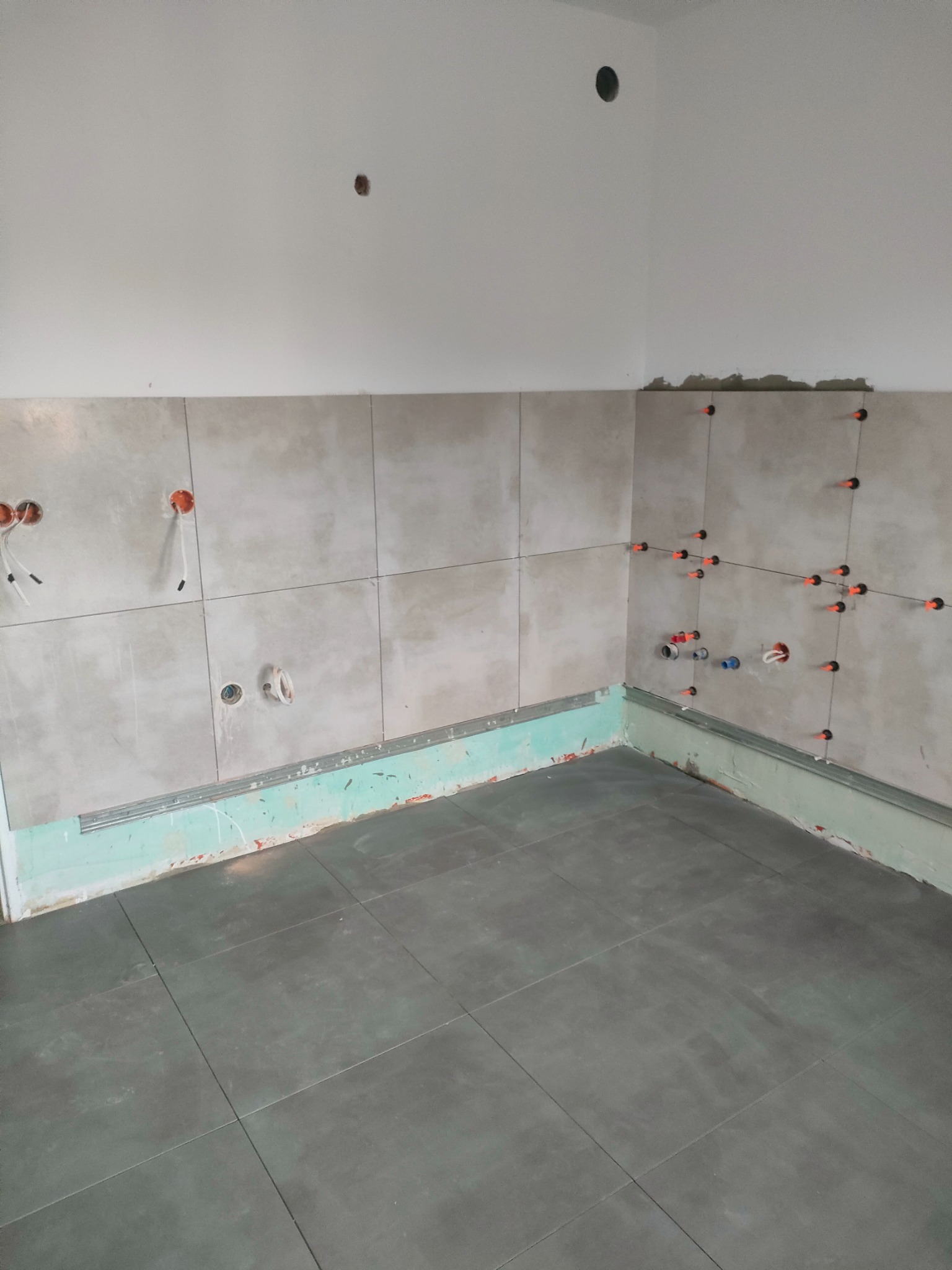 Fragment łazienki w trakcie prac: szare płytki podłogowe i ścienne, system poziomowania płytek, widoczne instalacje elektryczne i hydrauliczne.